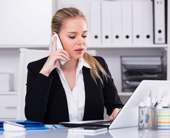 Smiling woman talking on mobile phone and using laptop in office